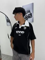 Chomba negra de manga corta con cuello polo blanco y ribetes blancos en los hombros y mangas. Presenta un parche bordado en el pecho izquierdo y un logo estampado en el centro.