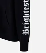 Saco tipo hoodie con cierre frontal de cremallera, color negro, corte boxy y capucha. Presenta estampa de letras en blanco en ambas mangas y un colgante de cruz metálico en el tirador de la cremallera principal.