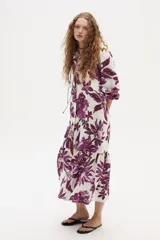 Vestido midi de lino con estampado floral grande en tonos morados sobre fondo blanco. Presenta cuello camisero, cierre frontal parcial con cordones negros y mangas tres cuartos abullonadas. La falda tiene un corte con volado en el ruedo.