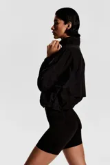 Campera negra de nylon repelente al agua y rompevientos con cuello elevado, cierre con protección para el mentón y bolsillos laterales con cierre. Modelo con hombros caídos, orificios de ventilación en las sisas y mangas murciélago con puños elásticos. Panel de ventilación en malla en la parte baja de la espalda y cordón para ajustar elástico con topes en el bajo. Forro de malla.