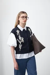 Chaleco tejido color azul marino con bordado aplicado de flores en hilo color crema. Presenta cuello camisero, manga corta y cierre frontal de botones.