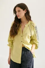 Camisa blanca de manga larga con bordados marrones en hombros y mangas, cuello en V y cierre frontal con botones.