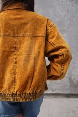 Campera estilo bomber o trucker de color mostaza/naranja quemado con efecto acid wash o desgastado. Presenta cuello camisero, cierre frontal y puños y cintura ajustados con elástico. Posee bolsillos frontales con solapa y botón.