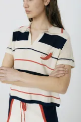 Remera polo tejida color crema con rayas horizontales en azul marino y rojo. Presenta cuello clásico, corte relajado y bordado de un ají rojo en el pecho.