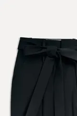 Pantalón negro de tiro alto confeccionado en mezcla de lana, con trabillas en la cintura, bolsillos laterales y falsos bolsillos de vivo en la espalda. Incluye cinturón de lazo en el mismo tejido y cierre frontal con cremallera, botón interior y ganchos metálicos.