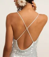 Vestido midi de malla con lentejuelas plateadas, escote recto y tirantes finos cruzados en la espalda. Presenta un forro corto que deja ver la transparencia de la malla con lentejuelas en la parte inferior, creando un bajo asimétrico.