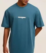 Remera relaxed azul petróleo de algodón, con cuello redondo en rib, manga corta y estampa localizada en relieve con la palabra "Eternal".