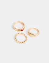 Set de tres anillos dorados de metal con diferentes diseños: uno liso, uno con corazones y uno con un corazón rojo.