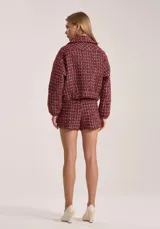 Chaqueta corta de tweed color borgoña con detalles en blanco. Presenta cuello redondo, cierre frontal con cremallera dorada, bolsillos delanteros y ribetes deshilachados.