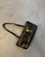 Cartera de hombro negra, de forma rectangular y acabado liso. Presenta detalles de hebillas plateadas y tiras decorativas en el frente, junto con un dije colgante en forma de llave dorada.