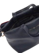 Cartera tipo tote de cuero sintético granulado color azul marino, con dos asas superiores y correa cruzada ajustable con cinta tejida a rayas azules y rojas. Presenta un cierre de cremallera principal y logo metálico de Tommy Hilfiger en el frente.