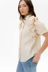 Camisa de lino color lila con volados en el cuello y en las mangas cortas abullonadas. Presenta un diseño de corte recto con cierre frontal de botones.