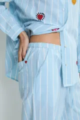 Conjunto de pijama celeste a rayas verticales blancas, compuesto por una camisa de manga larga con cuello mao y botones, y un pantalón de corte recto. La camisa y el pantalón tienen bordados de corazones, soles y otros motivos.
