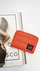 Billetera corta con cierre alrededor y tapa, confeccionada en tela capitoneada. Incluye varias reparticiones con tarjeteros y bolsillos internos, y una correa de mano desmontable. Disponible en negro, rojo y dorado.