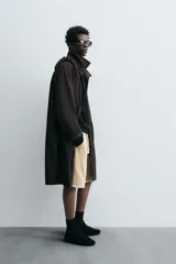 Trench relaxed fit negro confeccionado en tejido técnico ligeramente acolchado, con cuello subido y capucha desmontable. Tiene manga larga, bolsillos de vivo en pecho y de solapa en cadera, y abertura en el centro de la espalda. El cierre frontal es con cremallera oculta por solapa con botones a presión.