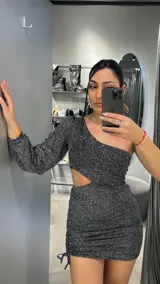 Vestido corto negro con brillos plateados, de un solo hombro y manga larga. Tiene un corte en la cintura y frunces laterales con cordones ajustables.