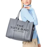 Bolso tote grande de cuero color gris con el texto "THE TOTE BAG MARC JACOBS" estampado en negro en el frente. Tiene doble asa de mano y cierre con broche a presión.
