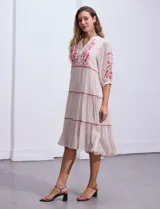 Vestido midi blanco con bordados florales azules en el escote, mangas y cintura. Tiene cuello en V, mangas 3/4 con botón y falda escalonada con detalles de ribetes azules.