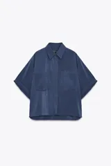 Camisa azul de cuello solapa y manga corta con dobladillo. Tiene dos bolsillos de parche en el pecho, bajo asimétrico con aberturas laterales y cierre frontal con botones ocultos.