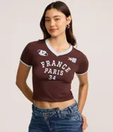 Remera femenina de media malla de algodón color marrón, con cuello en V y ribetes celestes en cuello y mangas. Presenta un estampado frontal con el lettering "France Paris 34" en celeste, junto con detalles deportivos como un logo "42" y una estrella con "Leurside".