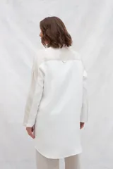 Camisa larga de lino color beige, con cuello camisero y cierre frontal con botones. Tiene mangas largas con puños abotonados, bolsillos laterales y una franja a contratono en la espalda y mangas.