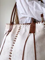 Bolso tipo tote bag de cuero color crudo con efecto craquelado y costuras de cuero marrón a contraste. Tiene doble asa de hombro de cuero marrón y cierre con cordones de cuero marrón.