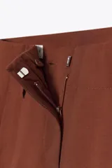 Pantalón marrón de tiro alto con bolsillos delanteros y falsos bolsillos traseros con tapeta. Presenta pliegues en la parte delantera y cierre frontal con cremallera y gancho metálico.