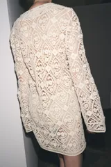 Vestido corto color crudo confeccionado en tejido tipo crochet con diseño geométrico floral. Tiene cuello solapa, abertura con botón en la parte superior y mangas largas acampanadas.