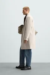Abrigo straight fit color beige confeccionado en tejido con lana. Cuello con solapas de muesca, manga larga acabada en puño con botones, bolsillos de vivo en cadera, detalle de bolsillo interior y bajo con abertura en centro espalda. Cierre frontal de botonadura.