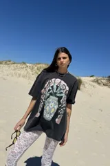 Remera de manga corta, color gris oscuro con efecto lavado, con un estampado frontal que incluye un diseño de planta en una maceta decorada, serpientes y texto. Se usa superpuesta a una remera de manga larga a rayas amarillas y blancas.