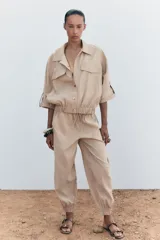 Camisa color beige de cuello solapa y manga corta con trabilla y botón. Tiene bolsillos delanteros de plastrón con solapa, bajo con elástico y cierre frontal con botones a presión.