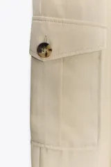 Pantalón color beige de tiro alto con bolsillos delanteros, bolsillos de plastrón con solapa laterales y falsos bolsillos de vivo en la espalda. Bajo acabado con trabilla y botón. Cierre frontal con cremallera y botón.