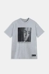 Buzo de manga corta color gris melange con cuello redondo. Estampado frontal en blanco y negro con el rostro de una persona con flequillo cubriendo parcialmente el rostro y el texto "nadie sabe lo que no se ve" en tipografía gótica estilizada. Incluye un parche con logos en el bajo.