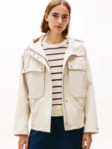 Campera beige de Tommy Hilfiger, con capucha con cordón ajustable y cierre frontal con cremallera y botones a presión. Tiene dos bolsillos delanteros tipo sobre con botón y logo metálico de Tommy Hilfiger en el puño.