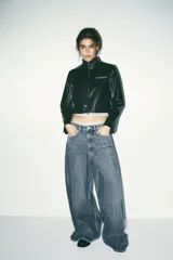 Cazadora corta negra de efecto piel, con cuello subido y manga larga. Tiene bolsillos delanteros con cremallera metálica y cierre frontal con cremallera.