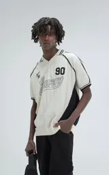 Chomba tejida de hilo color crudo con detalles en negro, de corte recto y manga corta. Presenta cuello tipo polo con rayas y bordado con el número 90 y la palabra 'Ravers'.