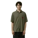 Camisa de manga corta con cuello camisero, patrón de pata de gallo en tonos verde oliva y negro. Posee un bolsillo frontal izquierdo con un parche bordado en rojo.