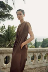 Vestido midi color marrón confeccionado en hilatura con mezcla de algodón y lino. Tiene cuello redondo, manga corta caída y detalle de pliegues en la cintura. Cierre en la espalda con abertura y botón.