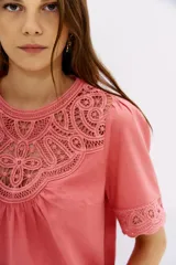 Camisa color salmón de corte recto, mangas cortas y cuello redondo. Presenta detalles de encaje en el escote y las mangas, y frunces en el frente y los hombros.