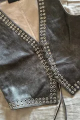 Chaleco negro de cuero con escote en V y cierre frontal con cordones. Presenta ojalillos metálicos plateados a lo largo de los bordes.