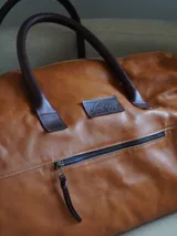 Bolso de viaje tipo weekender de cuero color marrón, con doble asa corta y correa larga desmontable. Cuenta con cierre superior, bolsillos internos y externos con cierre metálico. Forro interior de tela. Marca Sierra Mora.