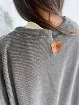 Buzo de cuello redondo en color gris con textura de punto, de corte holgado y hombros caídos. Incluye un detalle de parche decorativo en la parte posterior del cuello.