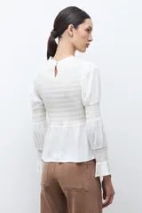 Blusa de color crudo con cuello alto fruncido y pechera con nido de abeja. Cuenta con mangas largas con detalles elásticos y puños con volados, y un bajo de corte peplum.