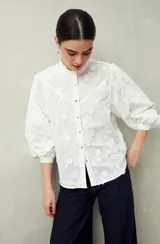 Camisa blanca con bordado floral en contraste, cuello mao, abotonadura frontal y mangas largas con frunces en los hombros.
