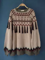 Sweater tejido con cuello redondo, mangas largas abullonadas y silueta holgada con tajos en los laterales. Presenta un diseño geométrico en tonos beige, marrón y verde, con flecos decorativos en la parte delantera.