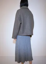Chaqueta color gris topo de tejido bouclé, con cuello camisero, cierre frontal con botones, dos bolsillos de parche con solapa y botón en el pecho, y mangas largas.