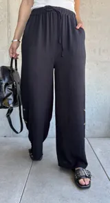 Pantalón negro de corte recto y pierna ancha, con cintura elástica ajustable con cordón y detalle de botones dorados en el lateral inferior.