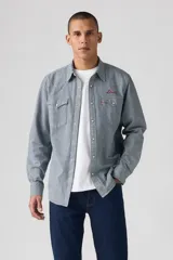 Camisa de manga larga a rayas verticales azules y blancas, con cuello americano, cierre frontal con botones a presión, dos bolsillos con solapa abotonada en el pecho y logo bordado en rojo sobre el bolsillo derecho.