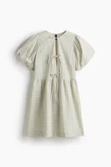 Vestido corto de algodón a cuadros en tonos beige y verde claro. Tiene cuello redondo con abertura y lazos, mangas cortas abullonadas y falda con leve vuelo.