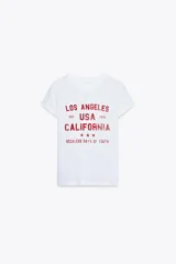 Camiseta blanca de algodón con efecto lavado, cuello redondo, manga corta con hombro caído y estampado frontal con texto 'Los Angeles USA California Reckless Days of Youth'.